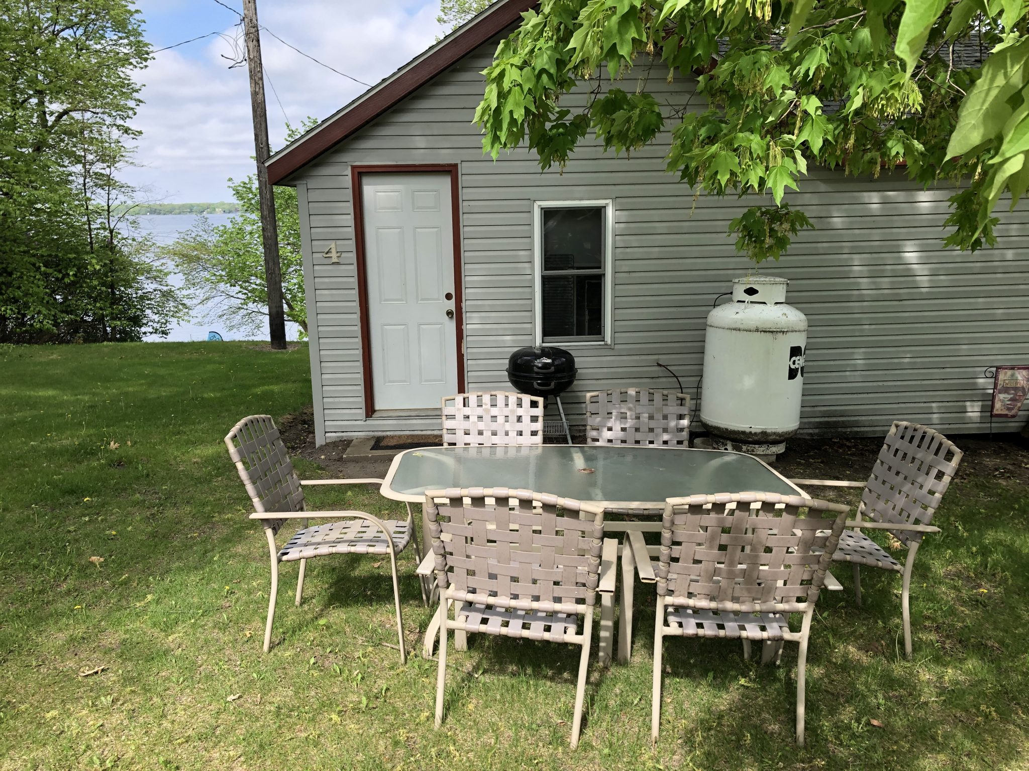 A small gray cabin with a white door and a glass-topped table with chairs on the lawn.